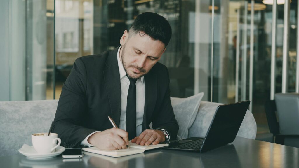 Professional businessman writing notes in a modern cafe setting, working on his laptop.